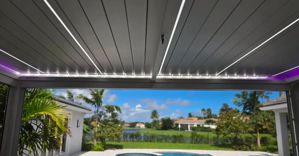 An upward interior view of a white louvered pergola ceiling with bright white linear LED strips integrated between the louvers, overlooking a backyard pool and palm trees.