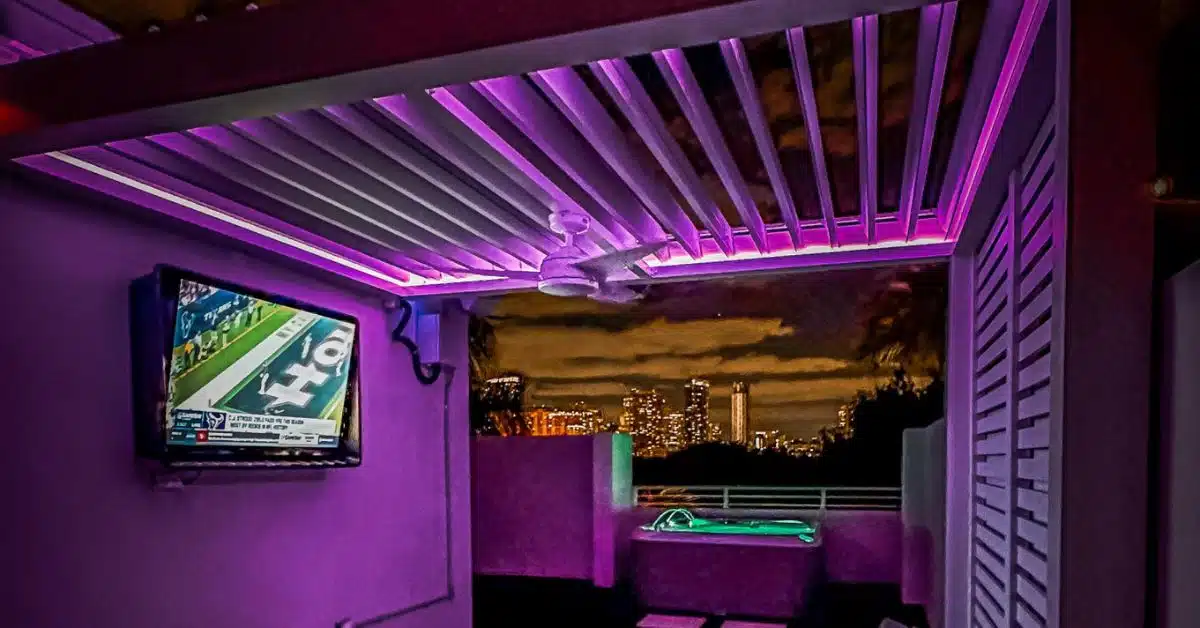 An interior nighttime view from a rooftop patio under a motorized louvered pergola featuring integrated linear RGB LED strip lighting displaying a purple color. The setup logicially integrates advanced technical displays, including a unique 'Freq.