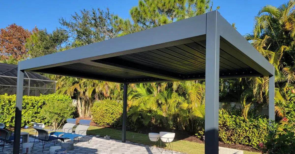 Freestanding Luxury Solid Roof Pavilion - enVision Pergola A freestanding dark grey solid roof pergola installed on a grey stone paver patio, surrounded by tropical landscaping and outdoor lounge chairs.