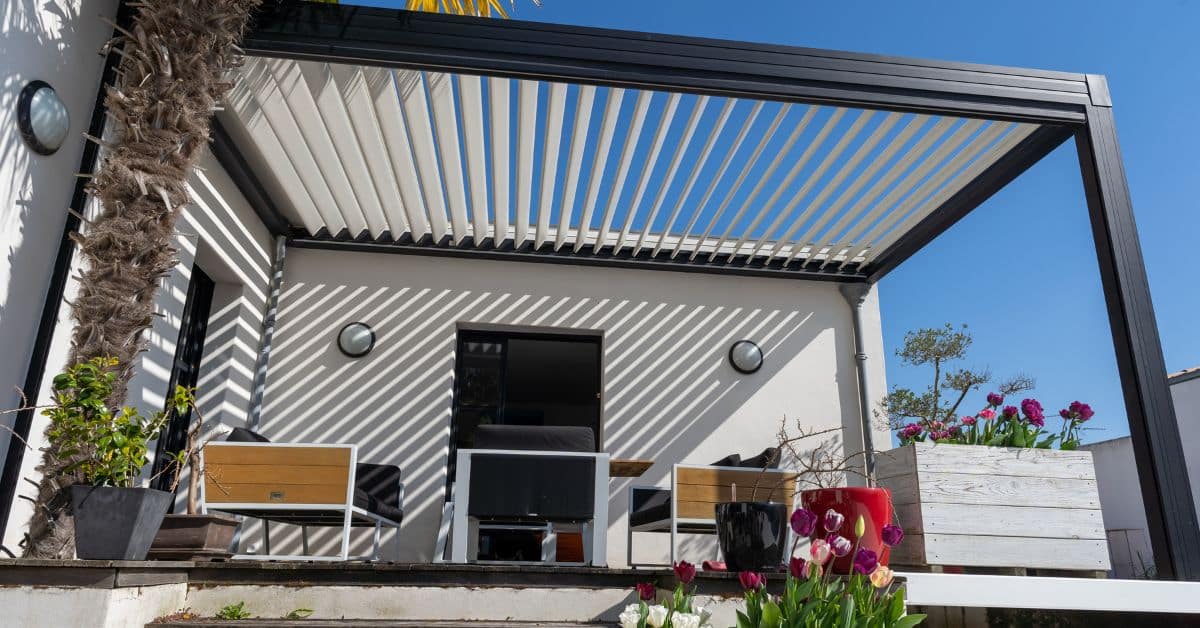 Adjustable Louvered Solid Roof System - enVision Pergola A modern black aluminum pergola with white louvered solid roof panels partially tilted to create a striking shadow pattern on the house wall.