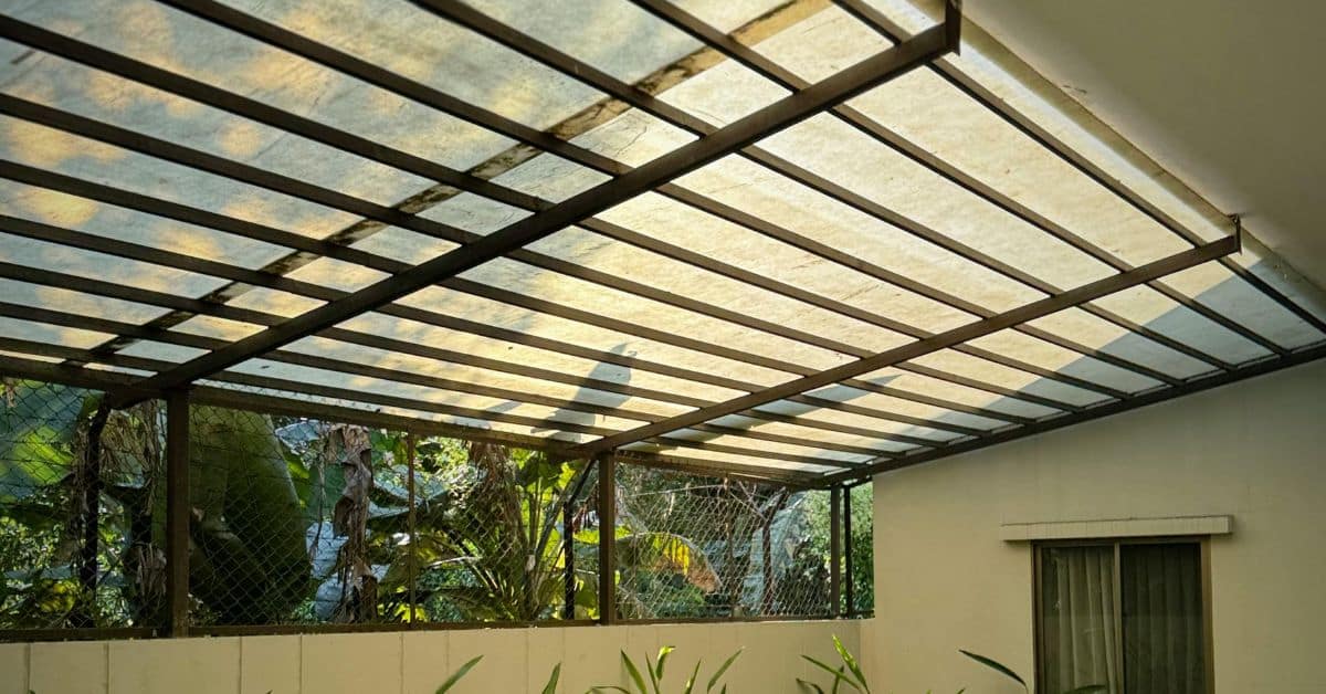 - enVision Pergola A wide-angle shot of an outdoor patio covered by a translucent polycarbonate solid roof pergola with a brown metal frame and internal support beams.