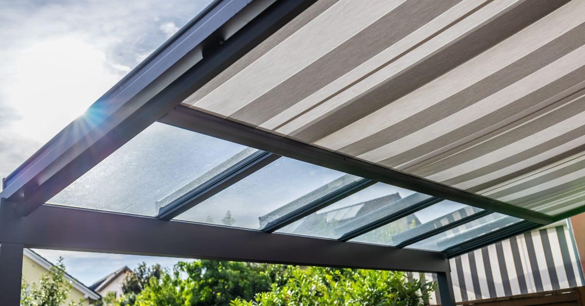 Solid Roof Pergola with Retractable Canopy A low-angle shot looking up at a modern dark grey aluminum pergola frame with integrated retractable patterned fabric panels and clear, solid glass or polycarbonate panels, set against a bright, partly cloudy sky.