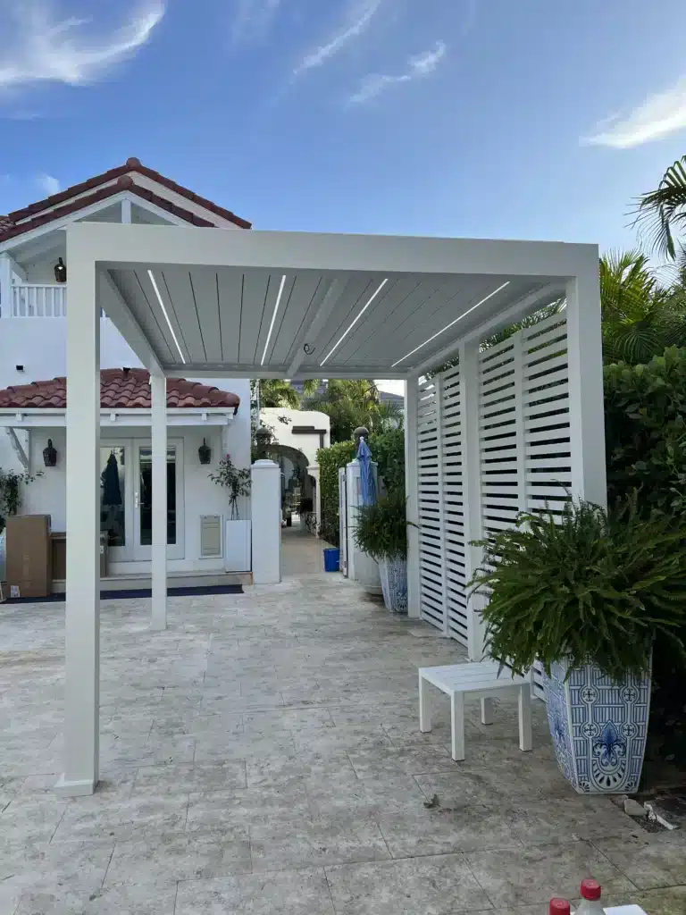White outdoor pergola over a stone patio, with a white slatted fence and large blue-and-white planters beside a white house with a red-tile roof.