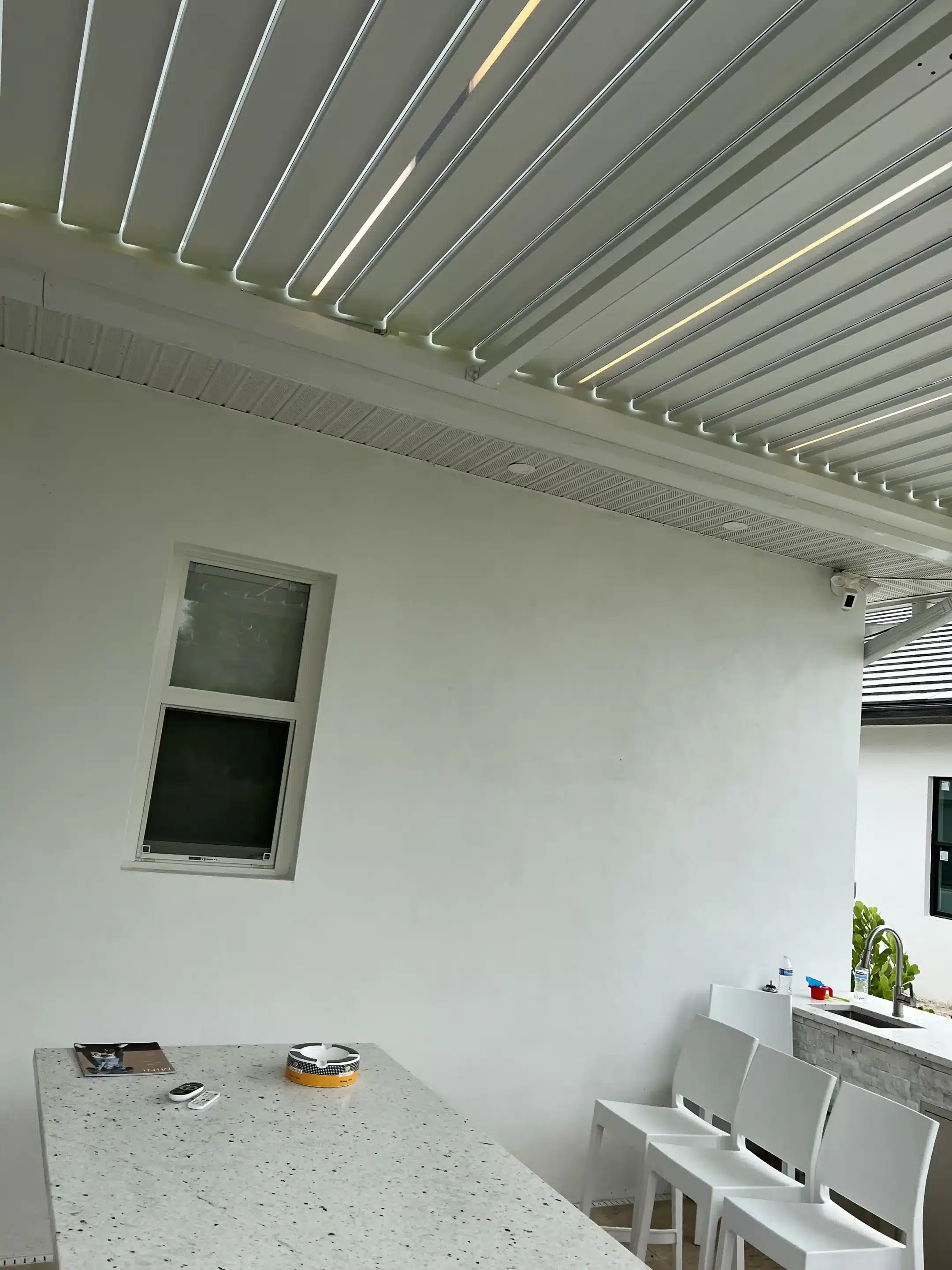 Modern kitchen/bar area with a speckled white island and white stools under a slatted metal ceiling, window on the left, and a sink area on the right.