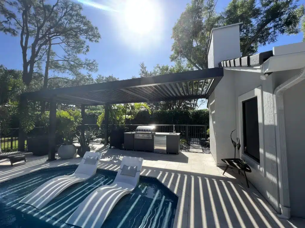 Backyard pool area with white lounge chairs, a metal pergola, and a modern outdoor grill under a bright sunny sky.