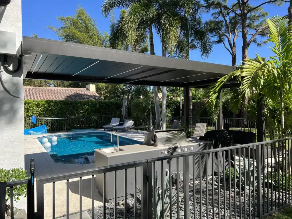Modern backyard pool area with a gray covered patio, lounge chairs, and palm trees under a blue sky, viewed from behind a fence.