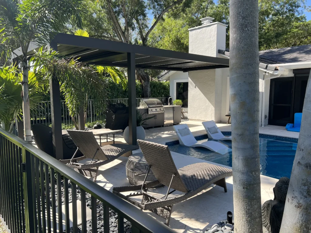 Poolside backyard with a dark pergola, white house, and a stainless grill under shade, surrounded by tropical plants and a railing nearby.