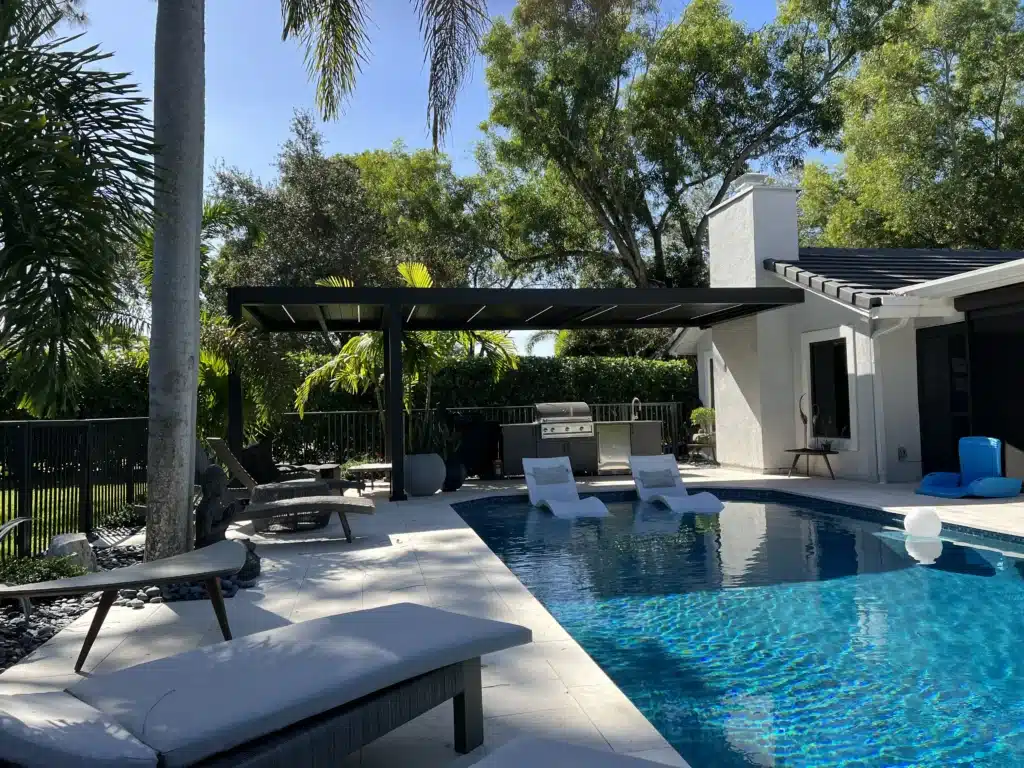 Outdoor pool area with white patio and modern black pergola, surrounded by palm trees and lush greenery under a clear blue sky. A seating area with white lounge chairs lines the pool edge, and a built-in grill is visible in the background near a white house.