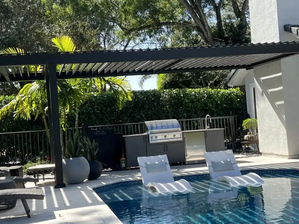 Poolside patio with white lounge chairs, a stainless grill, and a dark slatted pergola overhangs; lush green trees in the background.