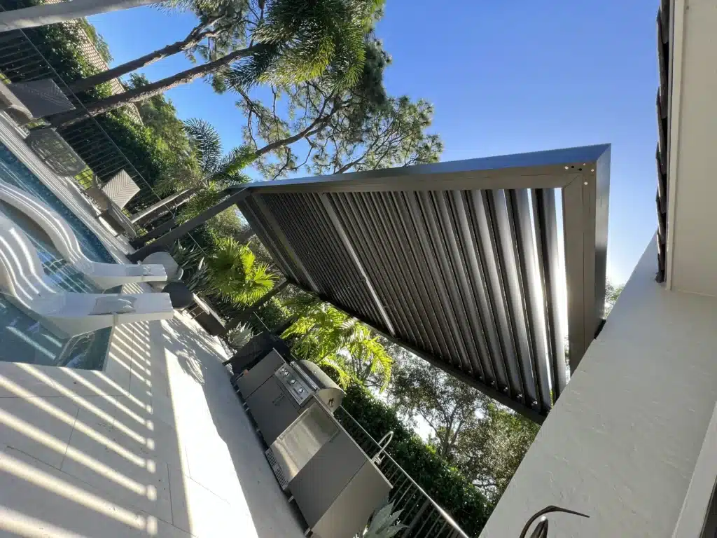Outdoor patio scene with white lounge chairs beside a pool, tall palm trees, and a dark slatted shade structure against a blue sky behind.