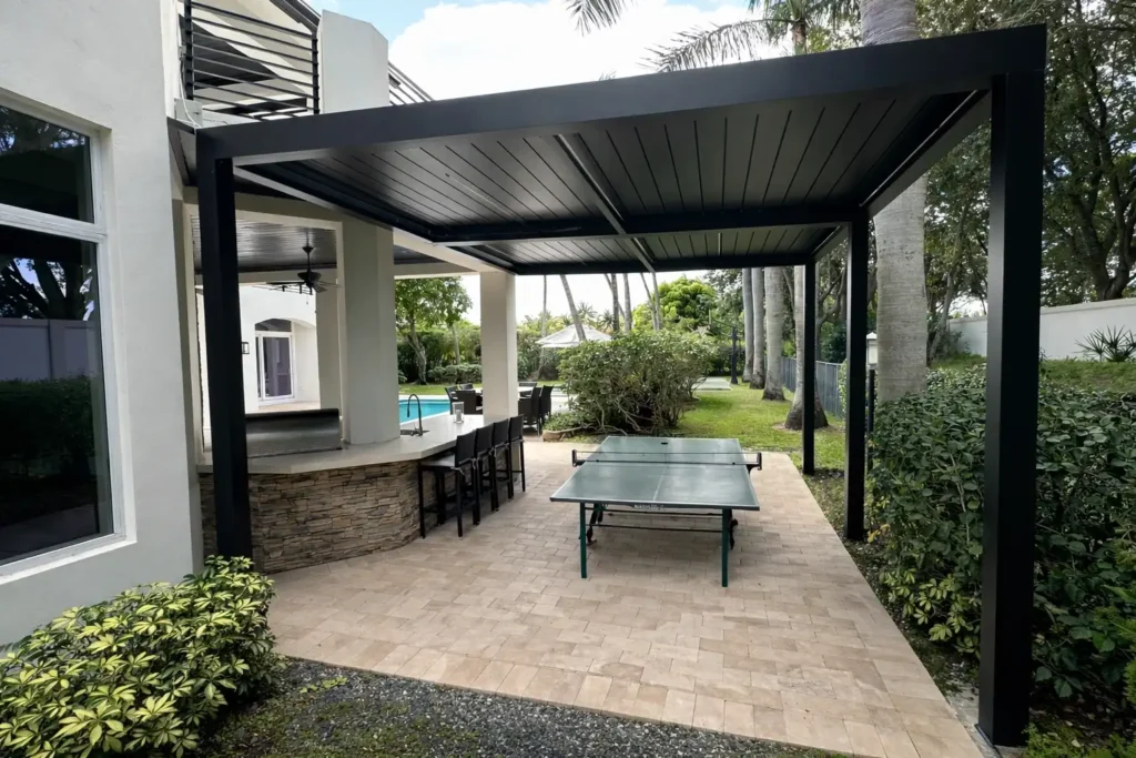 Covered outdoor patio with a dark metal pergola, a curved stone bar and bar stools, adjacent to a pool area.