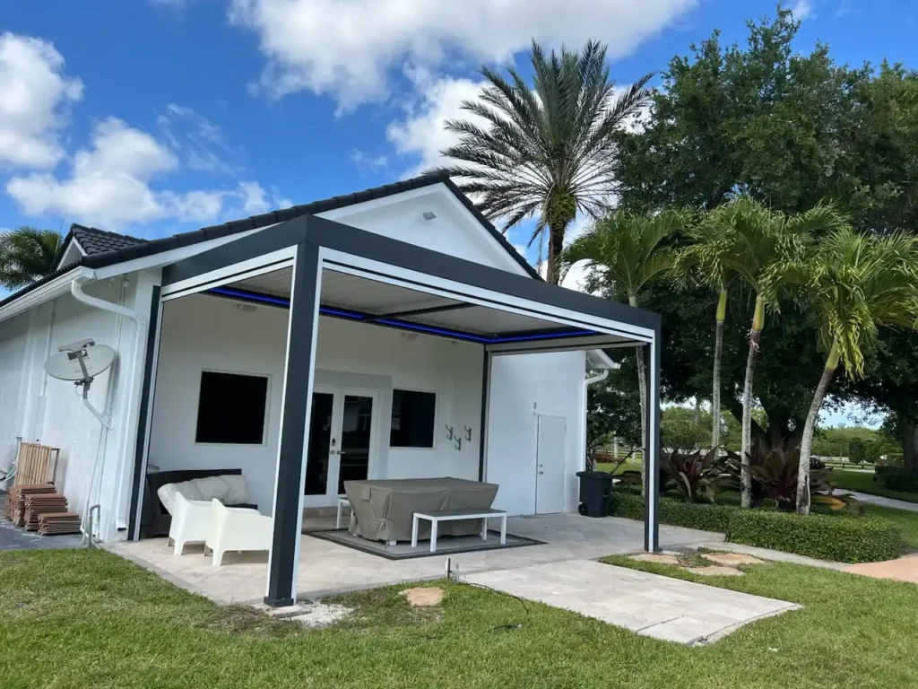 White single-story house with a dark-framed open patio cover, tiled patio, and light outdoor furniture under palm trees.