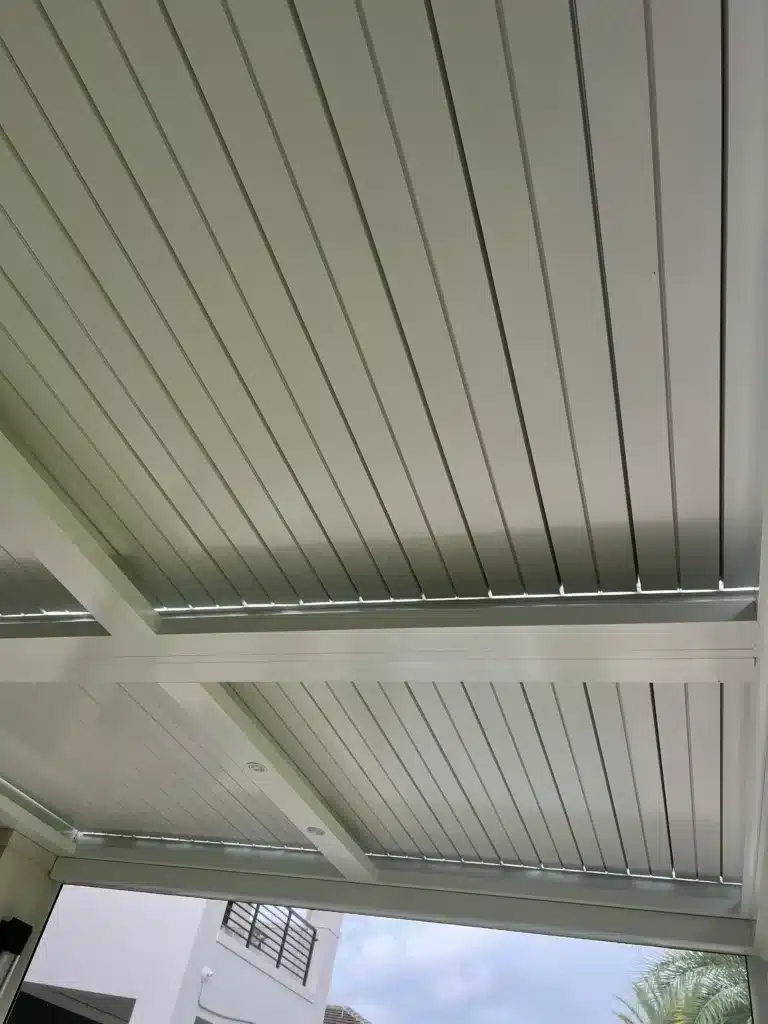 Interior view of a white slatted metal ceiling with supporting beams; outdoor scene visible through the open side showing a building balcony and palm tree