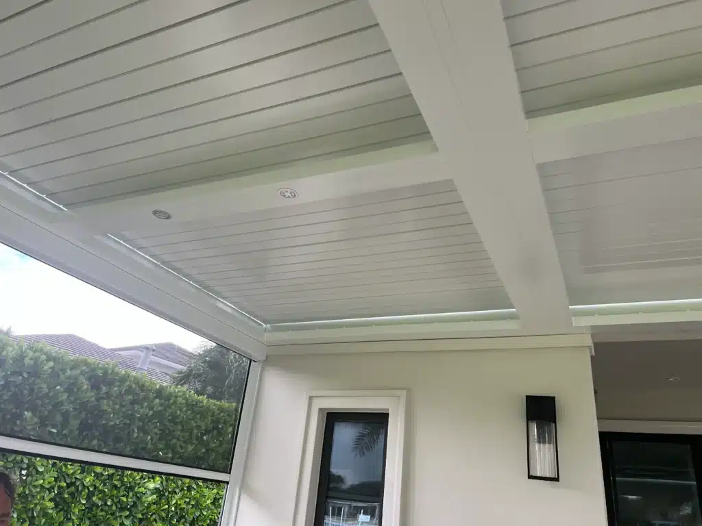 Modern covered patio area with white structural beams, glass walls, and a pale slatted ceiling; green hedges visible outside.