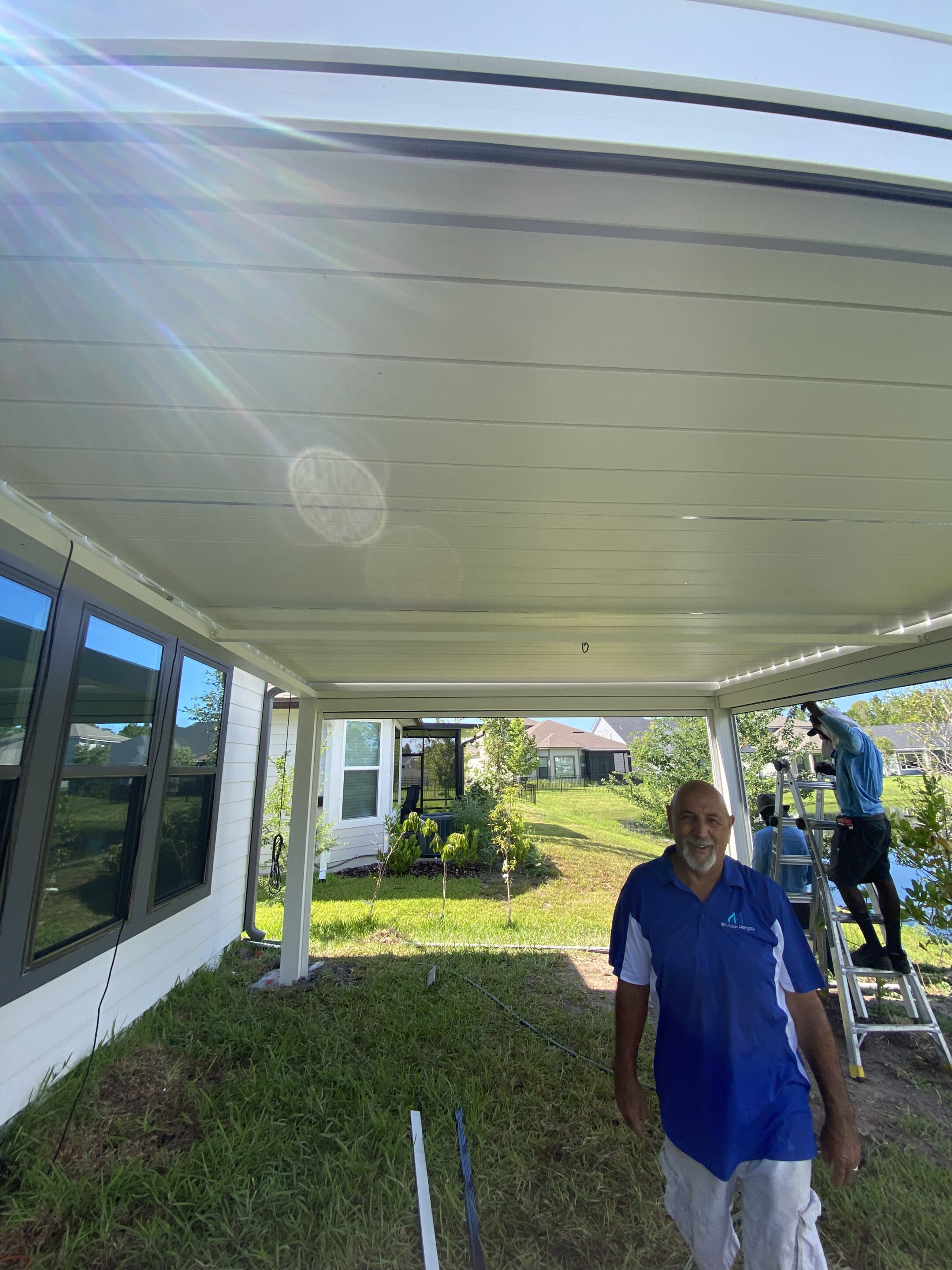 Two men work on a white patio cover: one on a ladder installing or inspecting, the other standing in front near the house and tools on the ground.