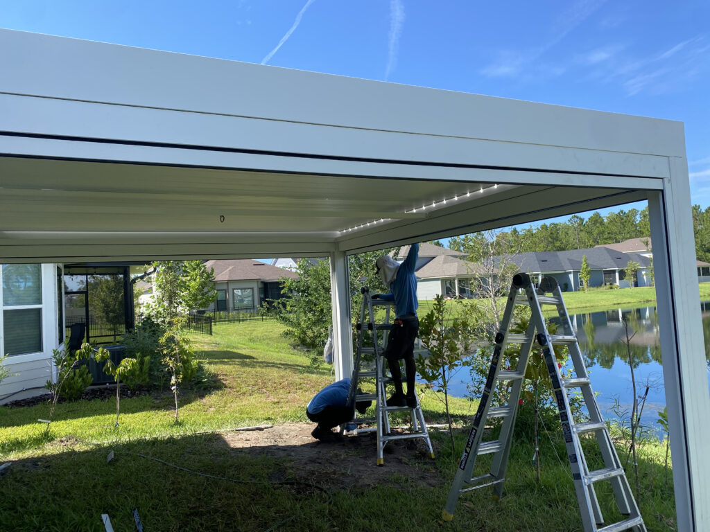 Construction workers on ladders installing a white aluminum patio cover in a backyard under a blue sky.