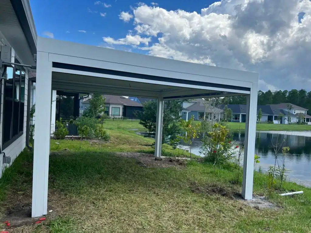 White metal pergola/carport frame over a grassy backyard with a pond and suburban houses beyond.