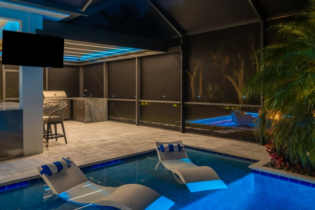 Nighttime poolside scene with blue-illuminated edges, lounge chairs, and a modern outdoor bar/kitchen along glass walls.