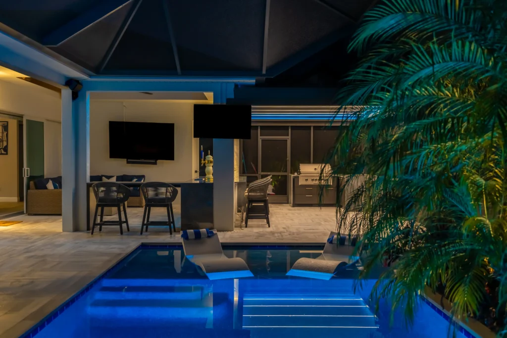 Nighttime outdoor patio with a rectangular pool, lounge chairs, a mounted flat-screen TV, and a grilling area under a covered patio, blue and warm lighting accents the scene.