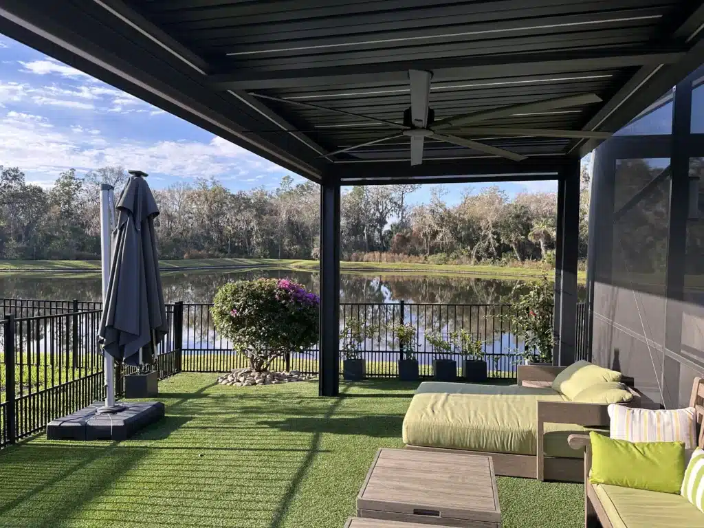 Outdoor covered patio with a large ceiling fan, black metal frame, and glass walls overlooking a lake and trees. A closed umbrella stands beside planters on artificial grass.