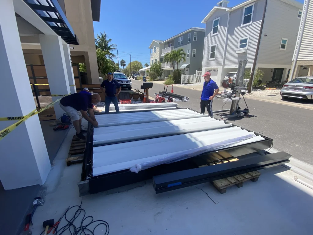 Construction workers in blue shirts unload long metal roofing panels on a residential street, with caution tape and tools nearby.