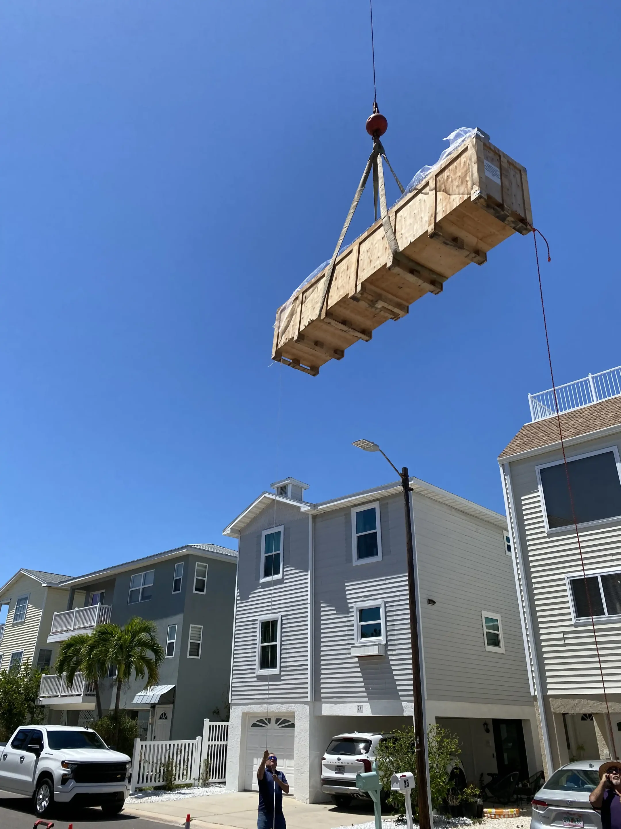 A wooden construction platform suspended by a crane being lowered over residential buildings against a clear blue sky.