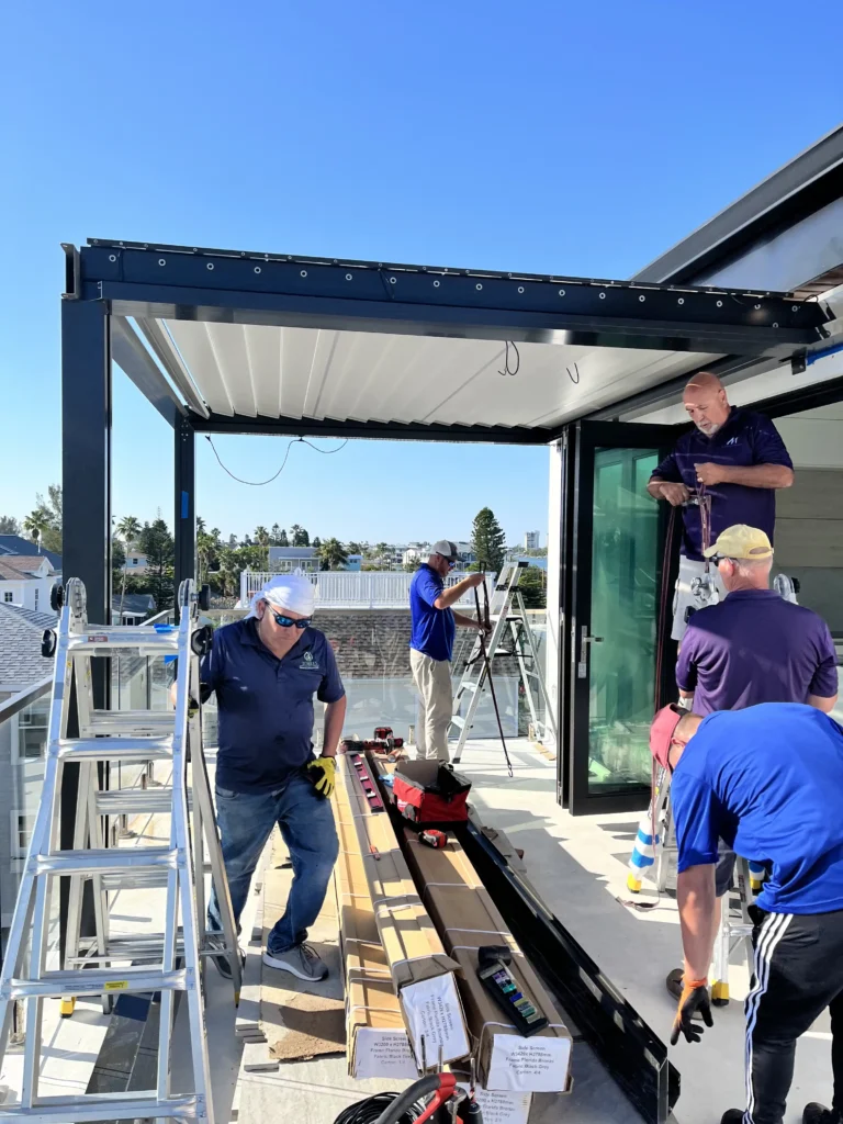 Construction workers install large glass railing panels on a rooftop balcony, with ladders and tools nearby against a clear blue sky.