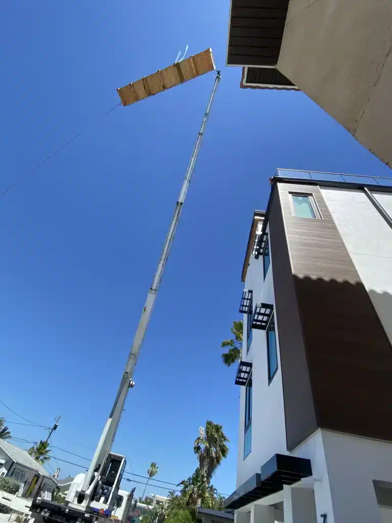 Crane lifting a long wooden beam near a modern building against a clear blue sky.