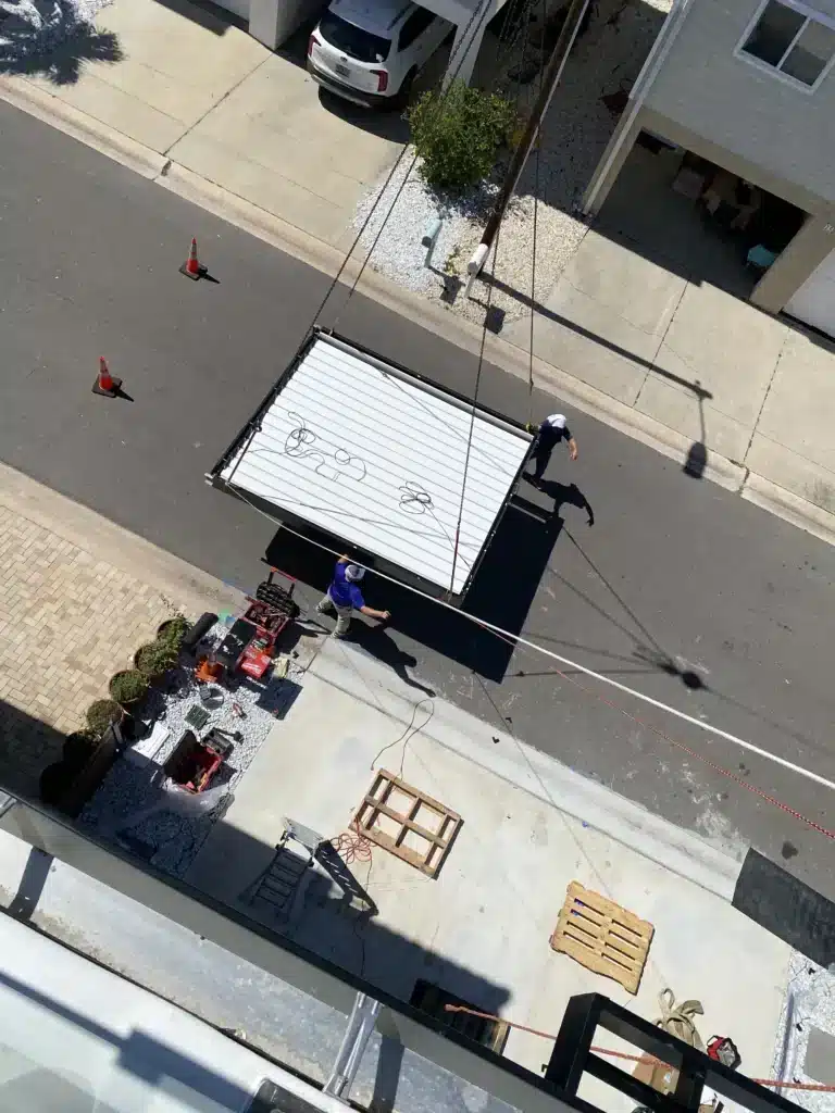 Aerial view of a construction scene: a trailer unloading onto a residential street with workers, cones, and a white car parked in the driveway.
