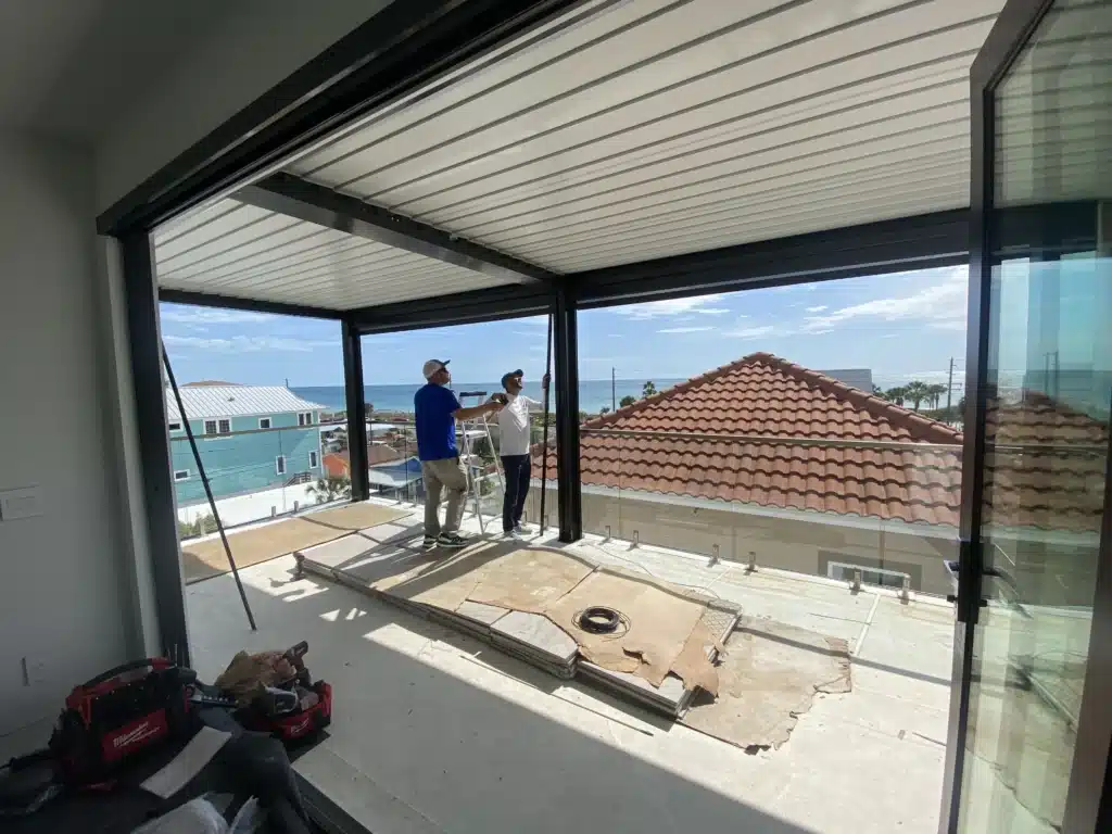 Two workers on an elevated balcony installing a glass railing, with the ocean and rooftops visible in the background.