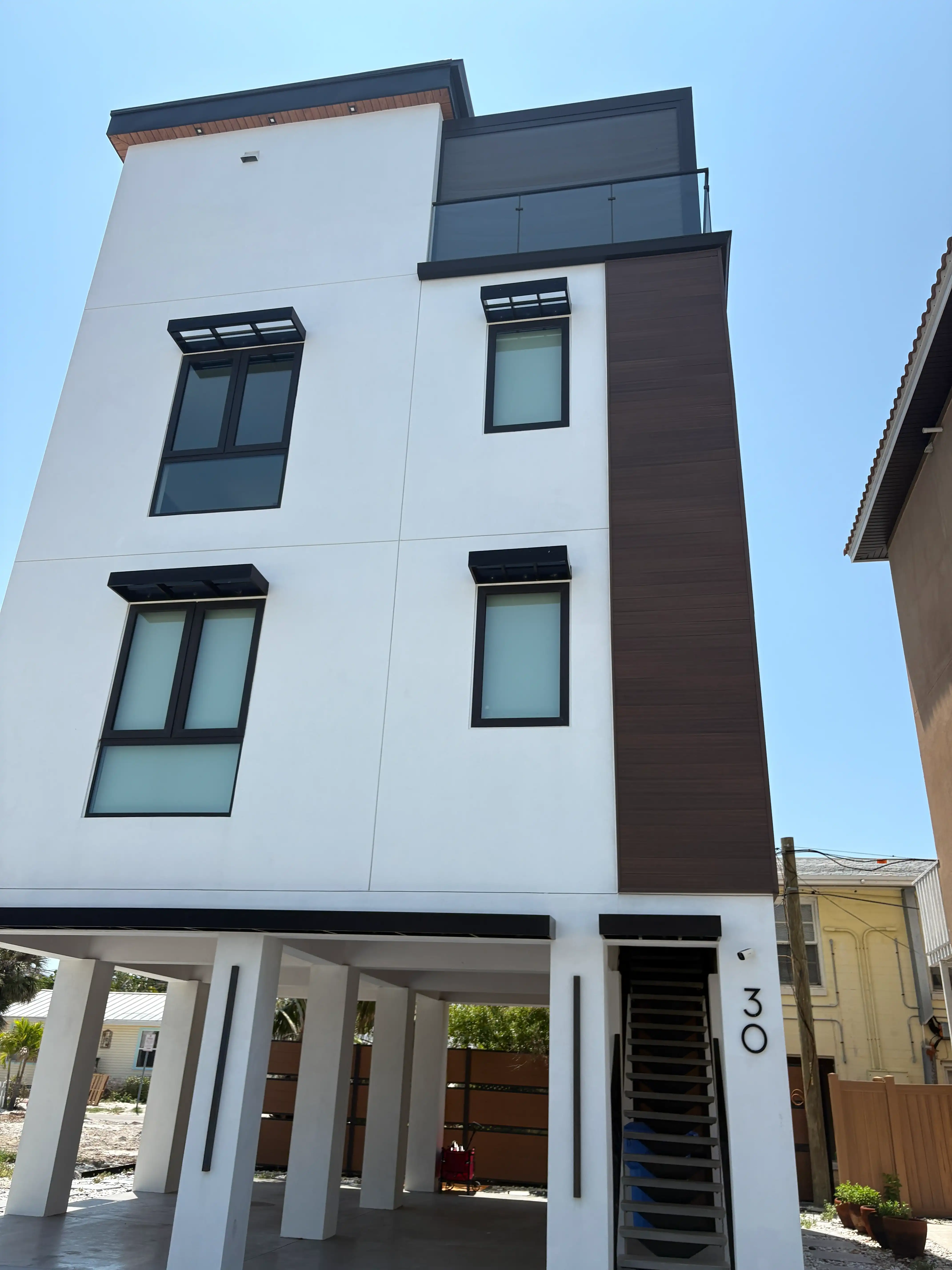 Modern white multi-story building with dark window frames, brown vertical panel, and an open ground-floor porch under a blue sky