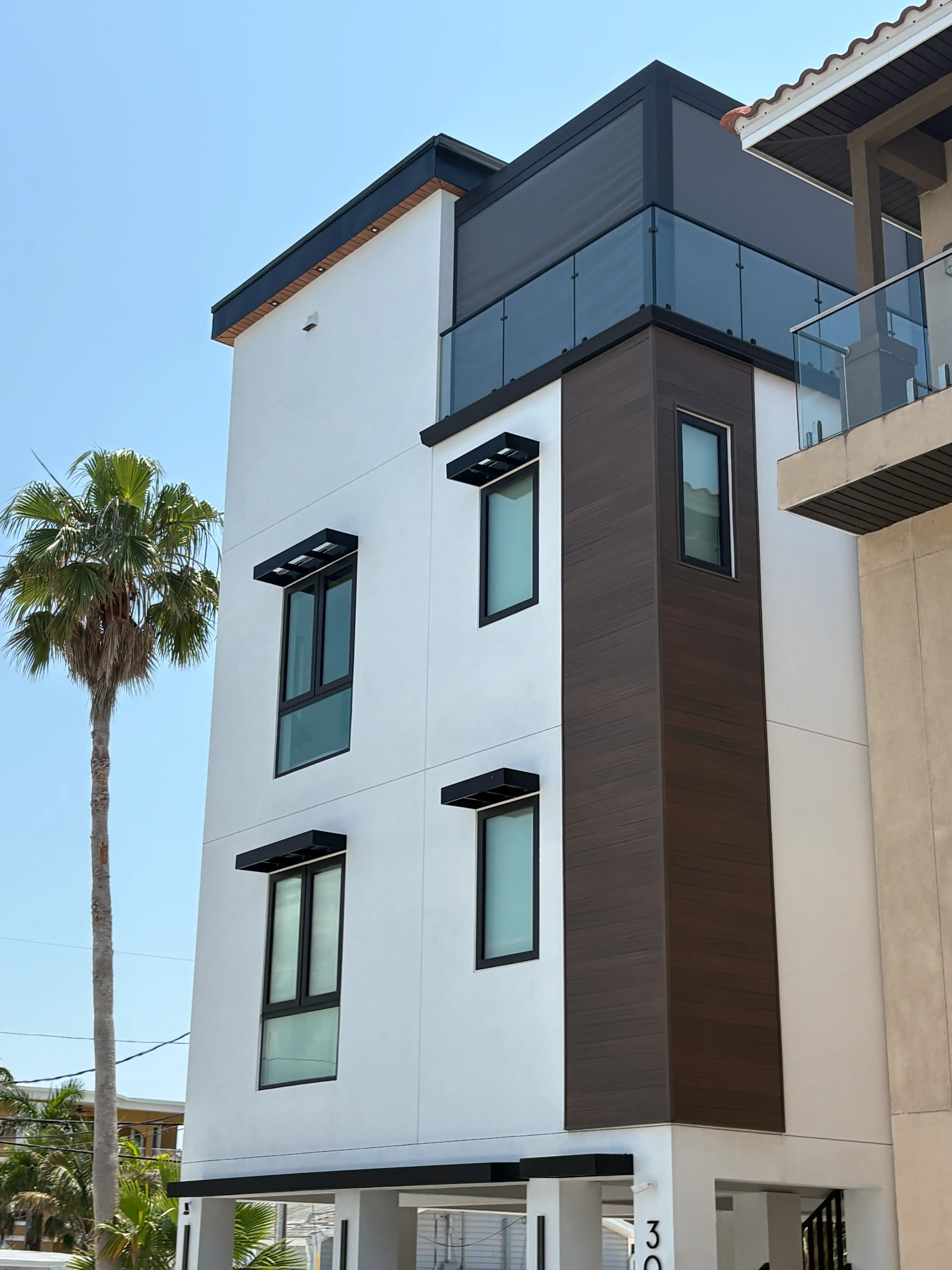 Modern white and brown apartment building with glass balcony railings against a clear blue sky, palm tree to the left.