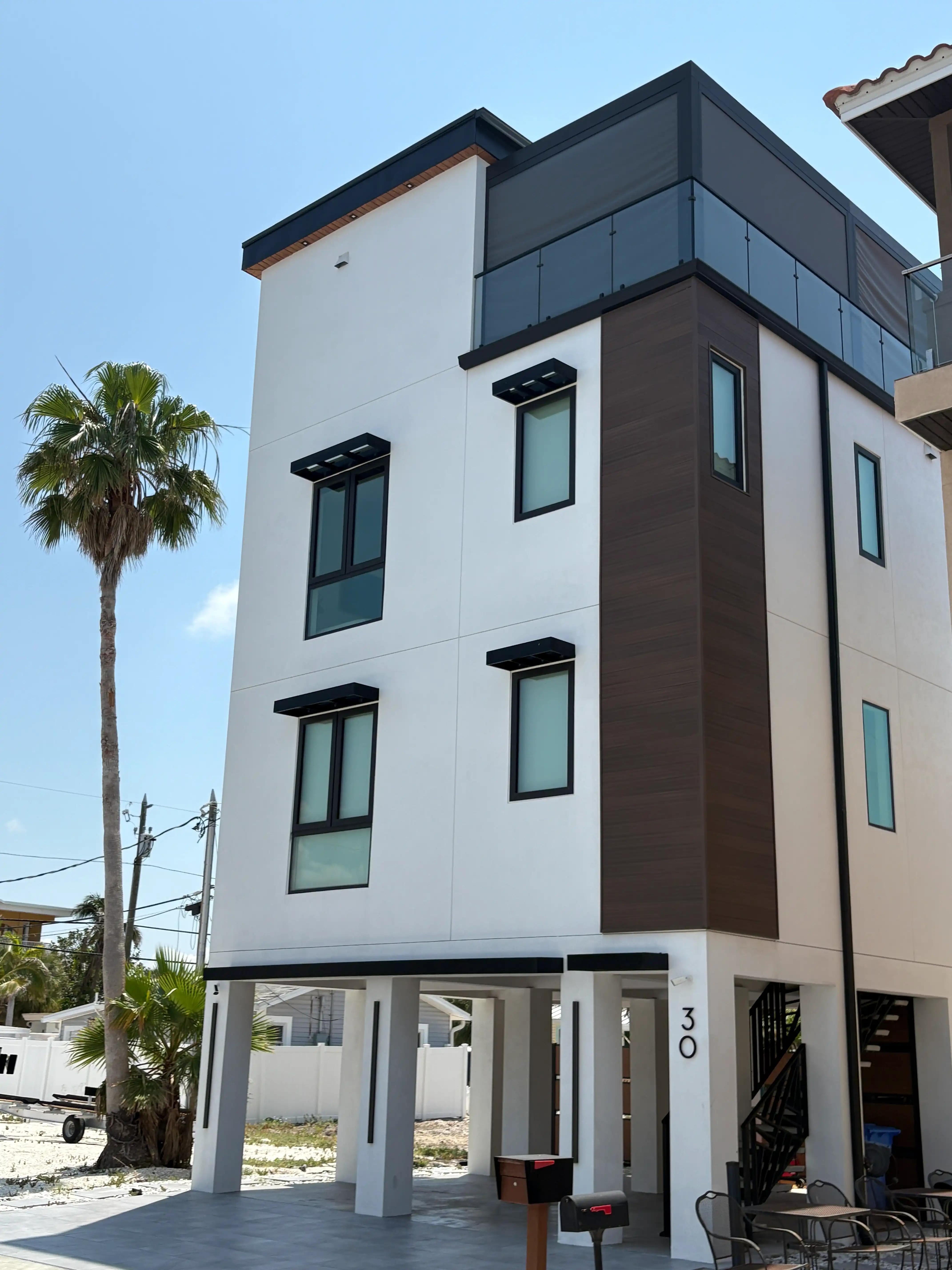 Modern white apartment building with dark vertical paneling, glass balcony railing on upper floor, and a palm tree nearby; ground-level parking area under the building.