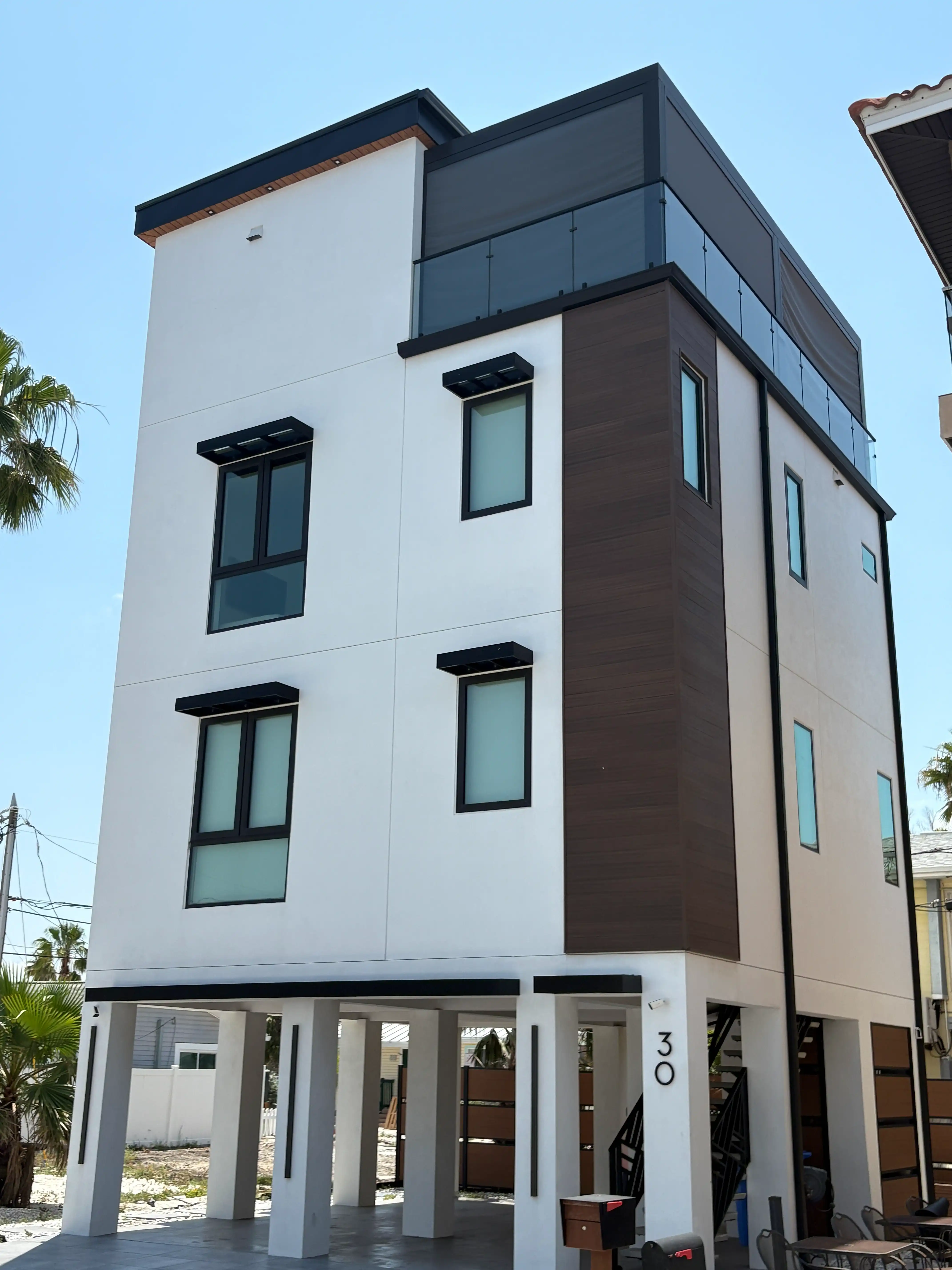Modern white apartment building with dark brown panels, glass balcony on the top floor, and a carport beneath.