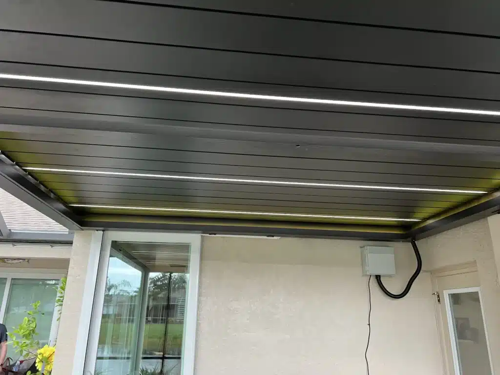 Outdoor patio area under a metal slatted ceiling with linear lights, sliding glass doors, and beige stucco walls.