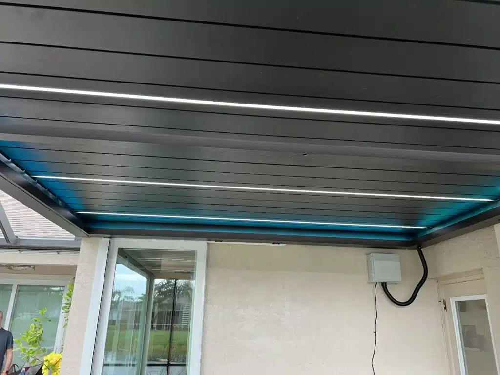 Outdoor patio with a metal slatted roof, linear LED lights, glass door panels, beige stucco walls, and hanging electrical cables.