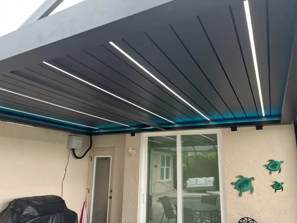 Covered outdoor patio ceiling with parallel slats and integrated linear LED lights, seen from below; sliding glass doors and beige stucco walls nearby.