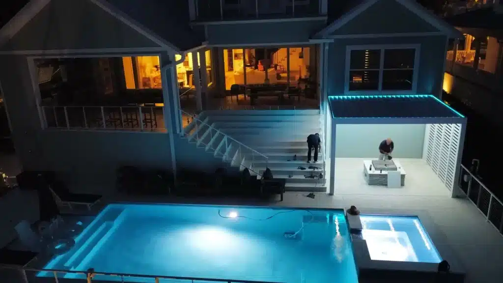 Aerial night view of a modern house with warm interior lighting, a covered patio, and a bright blue swimming pool below; two people on the deck.