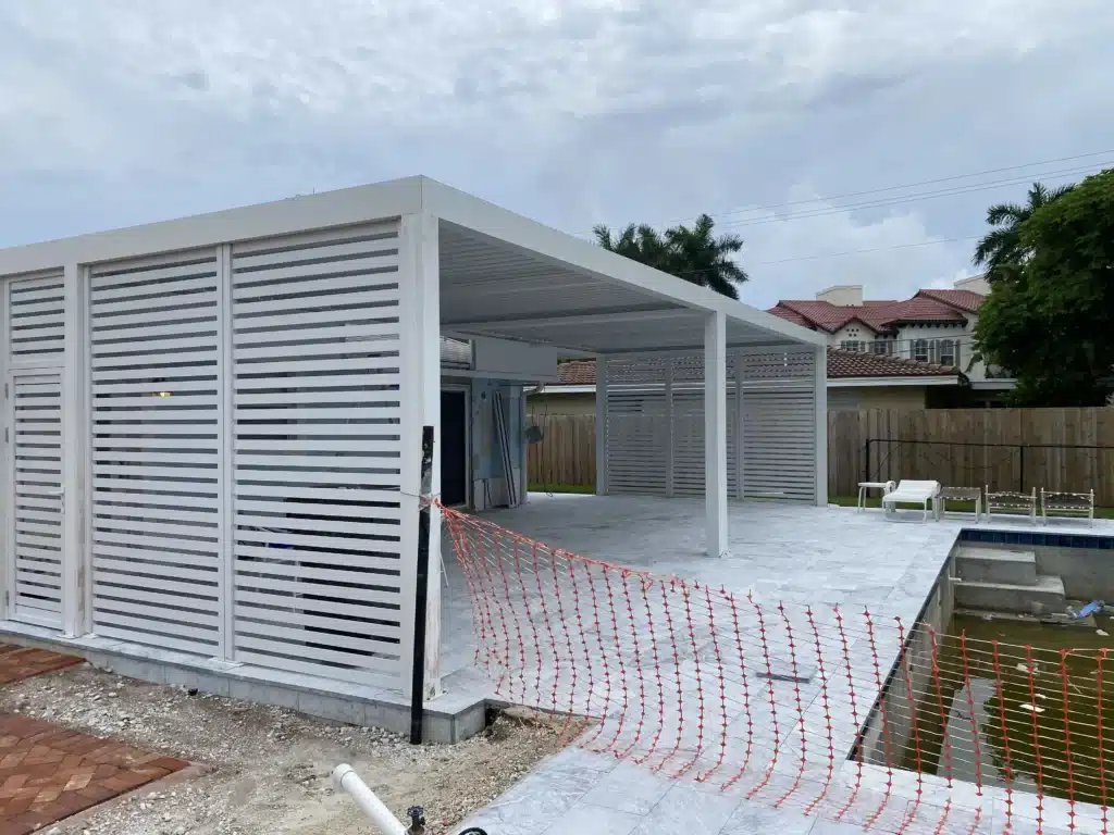 White outdoor pergola and privacy slat fencing under construction, with orange safety netting along the ground and a tiled patio area.