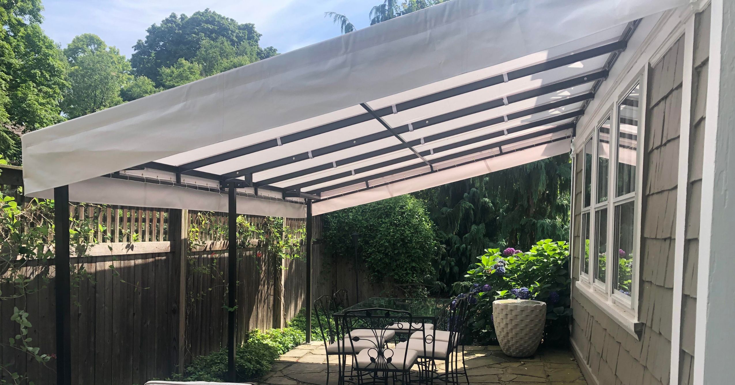 A dark metal pergola attached to a shingle-sided home, featuring a white weather-resistant fabric canopy over an outdoor dining table and chairs.