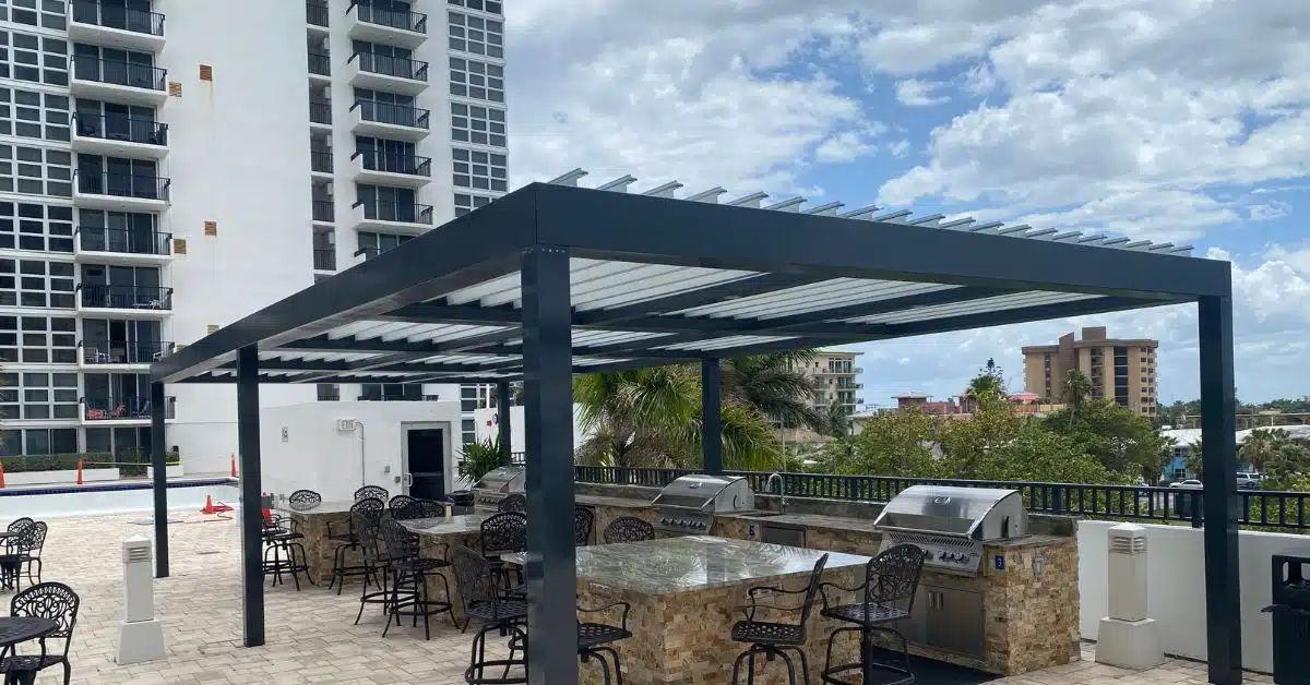 A high-end rooftop outdoor kitchen and bar area featuring stone-clad islands and stainless steel grills, sheltered by a large dark grey motorized louvered pergola system.