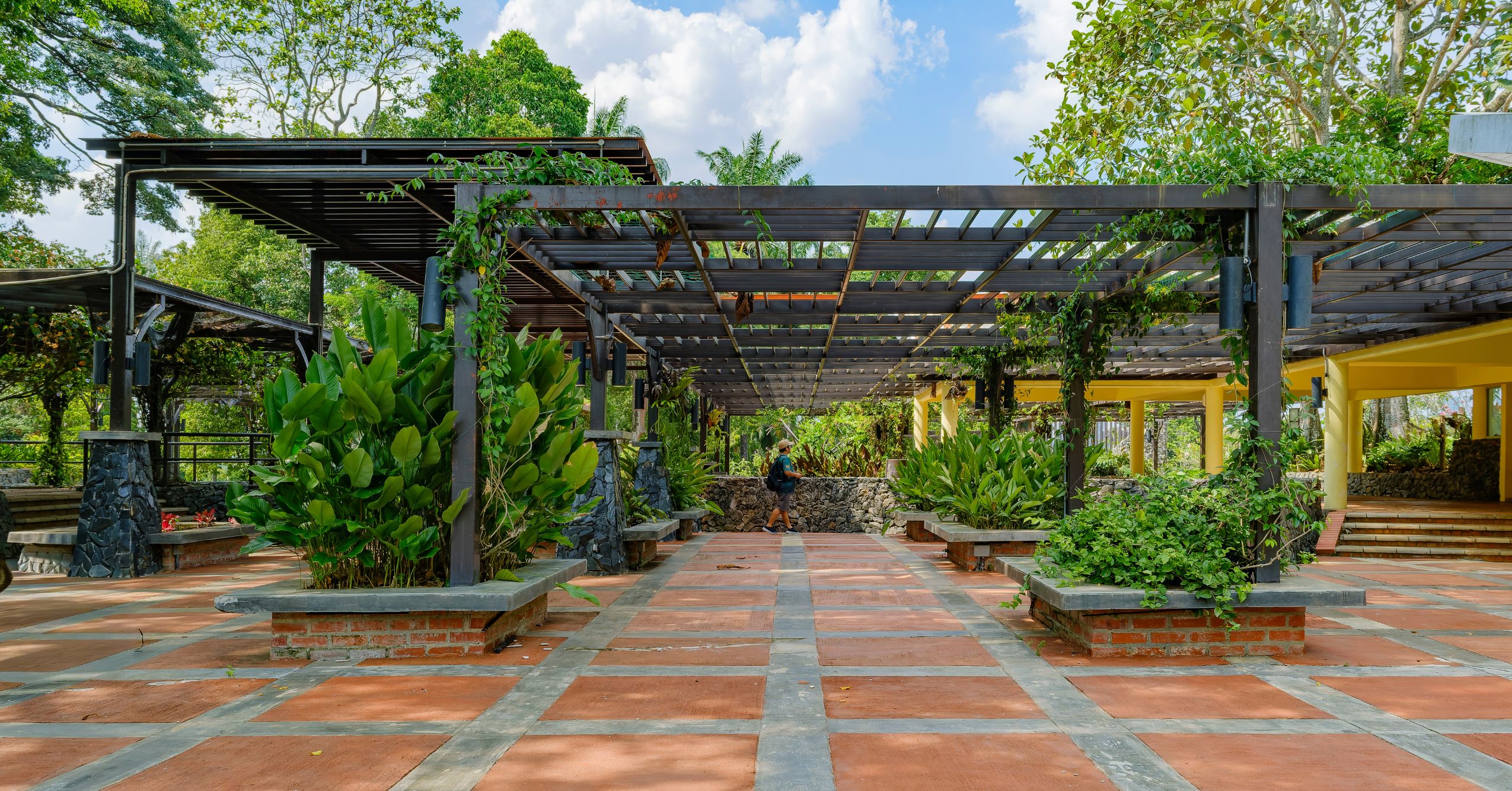 An extensive industrial-style metal pergola system spanning a public garden walkway with brick planters, lush tropical greenery, and stone pillars.