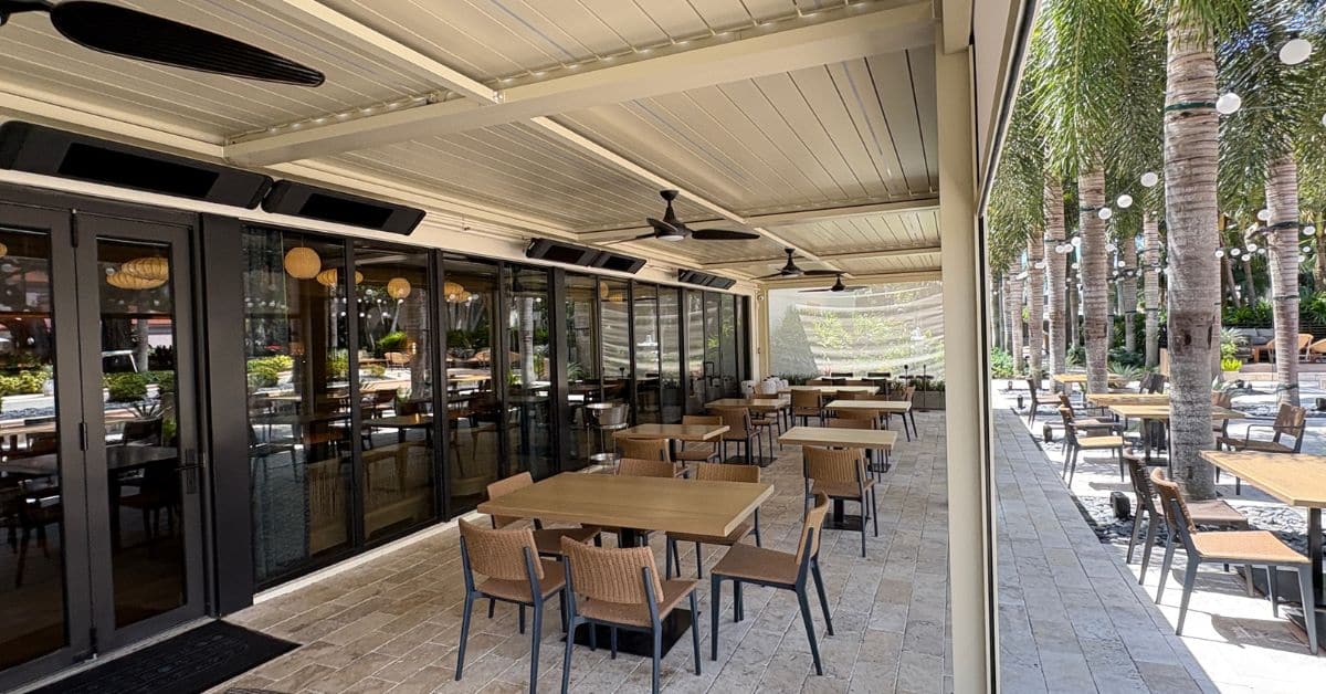 A wide view of a professional outdoor dining area under a large cream-colored automated pergola, featuring ceiling fans, infrared heaters, and glass sliding doors.