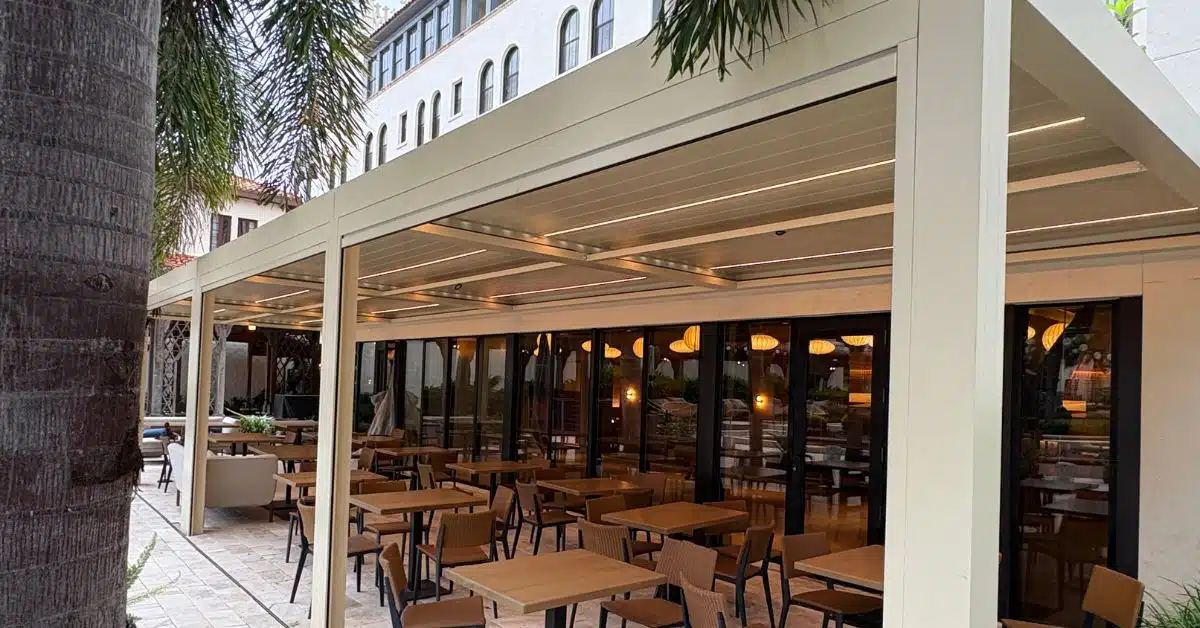 A minimalist matte black aluminum pergola casting strong geometric linear shadows against a neutral-toned textured stone wall in a modern backyard.