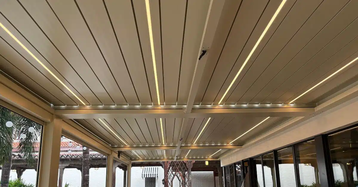 An upward view of a tan aluminum louvered roof system featuring integrated linear LED light strips between the panels for nighttime illumination.