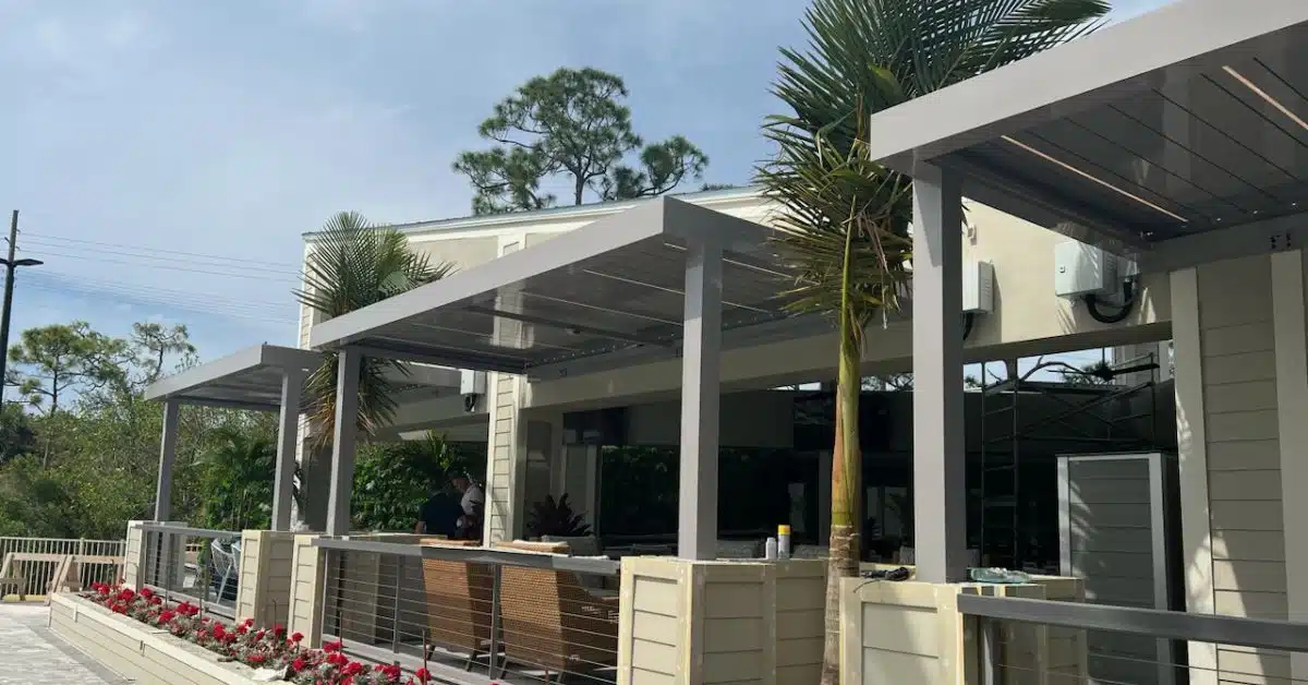 Modern Residential Outdoor Living with Motorized Louvers - enVision Pergola A grey residential motorized pergola system with integrated LED lighting installed over a luxury patio area with wicker seating and tropical landscaping.