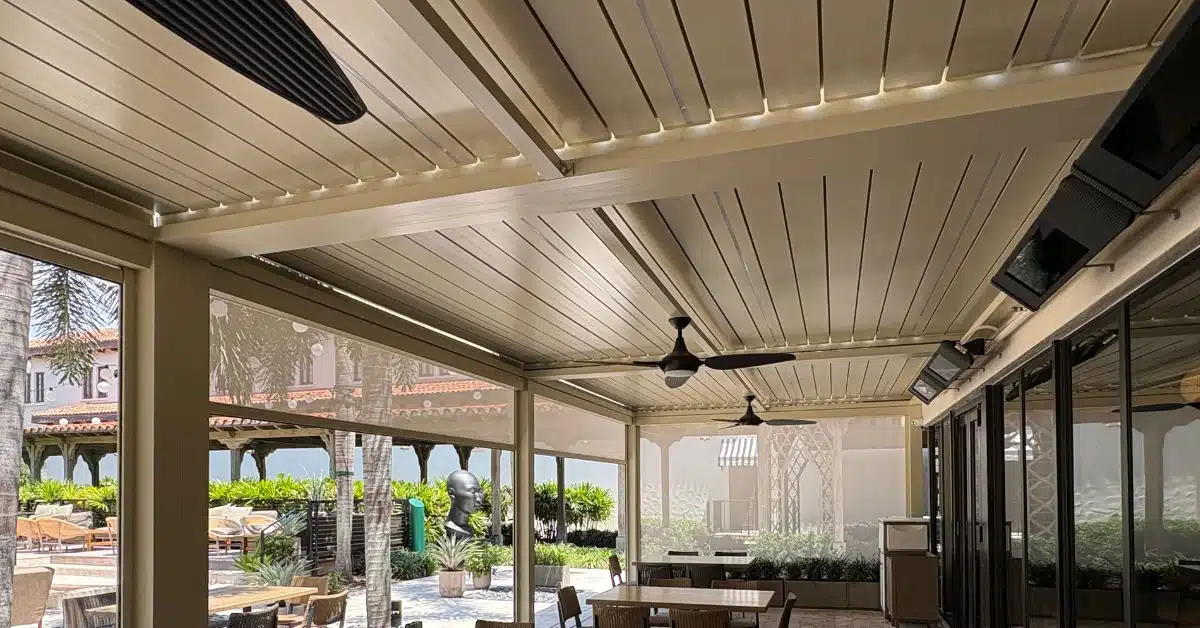 Motorized Pergola with Retractable Weather Screens - enVision Pergola An interior view looking out from under a motorized louvered pergola with semi-transparent retractable privacy screens lowered to block wind and sun.