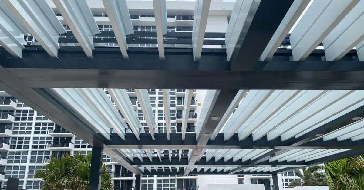 An upward view of a large white aluminum motorized louvered pergola with integrated lighting, supported by dark metal beams, set against a background of a multi-story white apartment building.