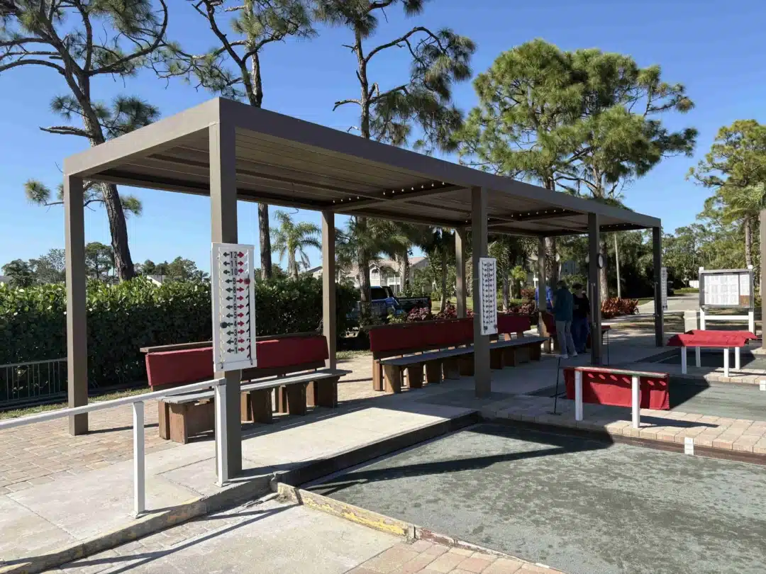 Outdoor event space under a brown pergola with wooden benches and scoreboards, palm trees and a clear blue sky in the background.