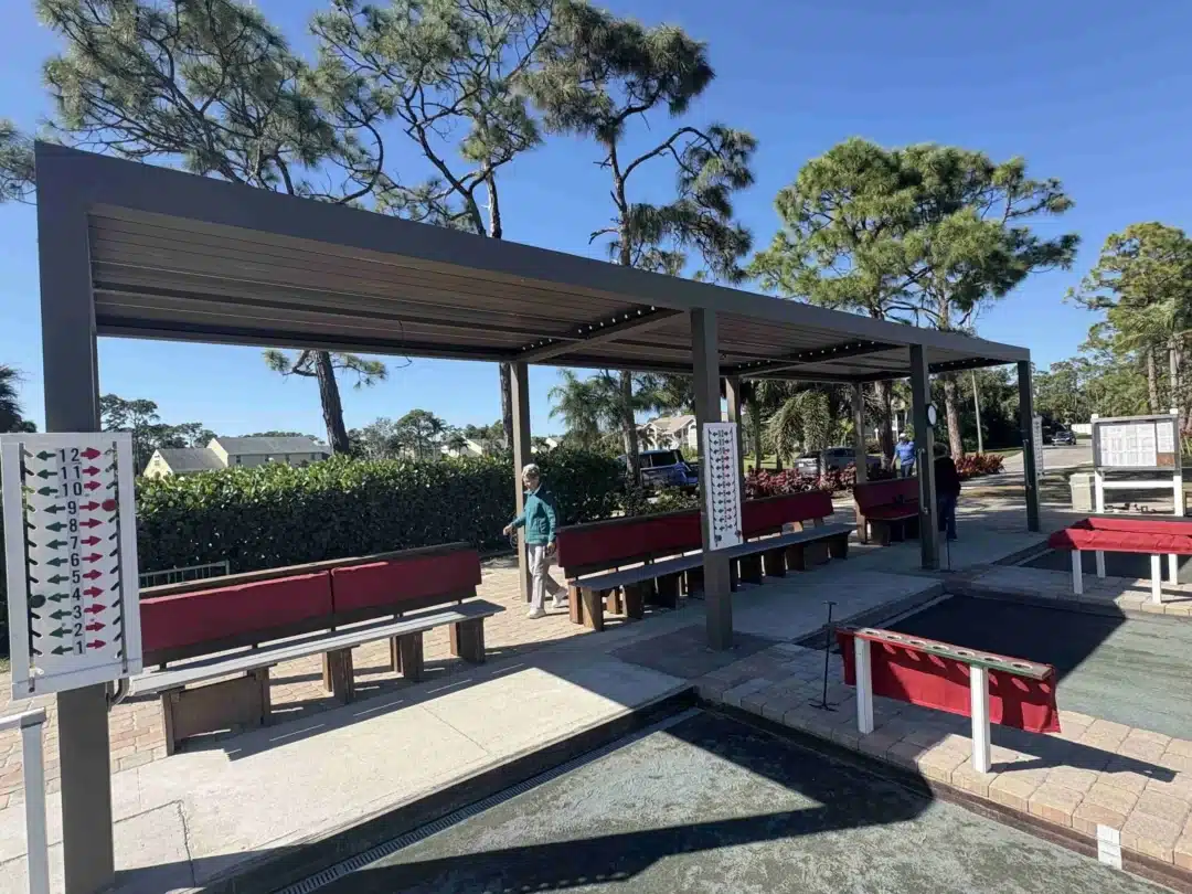 Outdoor seating under a gray metal pergola with red benches; a tall sign with numbered arrows on the left, trees and blue sky behind.